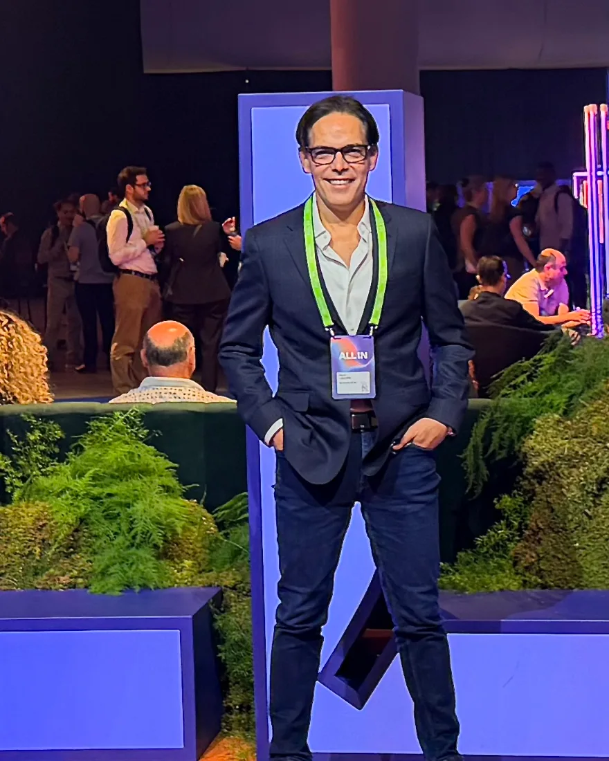 David Legendre standing and smiling, navy blazer, white shirt, jeans and glasses, ALL IN badge around his neck, in the networking area of the ALL IN conference in Montréal.